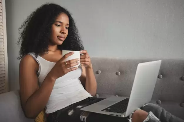 foto-de-lado-da-moda-jovem-atraente-de-pele-escura-em-jeans-rasgados-relaxando-no-sofa-com-o-computador-portatil-no-colo-bebendo-cafe-e-assistindo-a-serie-de-tv-favorita-online_343059-1584